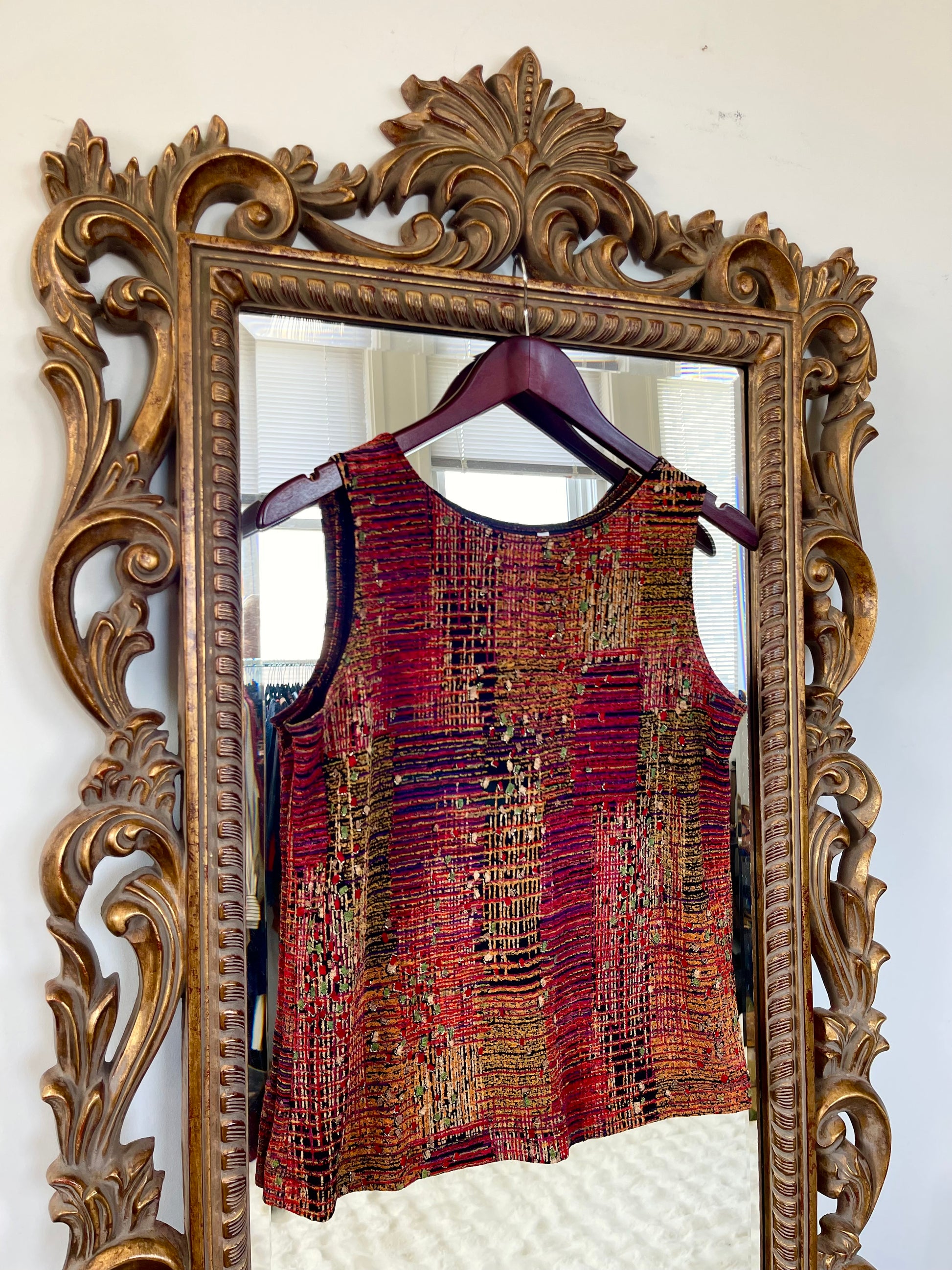 Decorative mirror with ornate frame reflecting a textured red garment on a hanger.